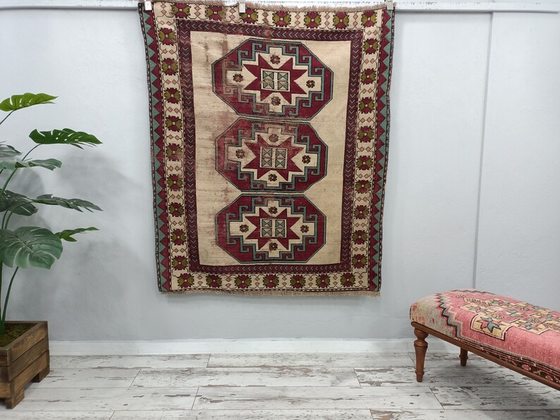 May include: A vintage Turkish rug with a cream background and three red, blue, and green geometric patterns. The rug is hanging on a white wall.