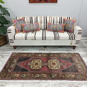 May include: A vintage pink and cream area rug with a dark brown center and intricate floral patterns. The rug is laid out on a white wood floor in front of a white sofa with striped cushions and colorful throw pillows.