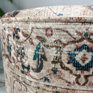 May include: A round, beige ottoman with a floral pattern in shades of brown, teal, and pink. The ottoman has a decorative trim around the top edge.