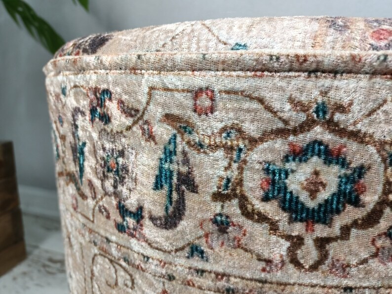 May include: A round, beige ottoman with a floral pattern in shades of brown, teal, and pink. The ottoman has a decorative trim around the top edge.