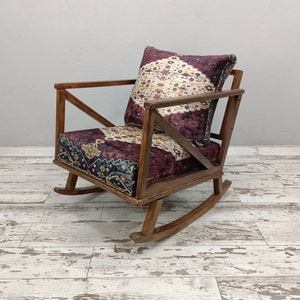 May include: Wooden rocking chair with a patterned cushion and backrest. The cushion and backrest feature a burgundy, cream, and navy blue design. The chair has a dark brown wooden frame and is set on a white and gray wooden floor.