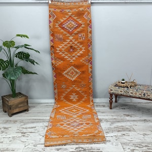 以下が含まれることがあります： 幾何学模様のオレンジと白の長いランナーラグ。ラグは下にフリンジがあり、白い壁にかけられています。
