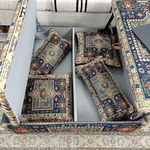 May include: A storage ottoman with a lift-top lid revealing a storage compartment. The ottoman is covered in a colorful patterned fabric with blue, red, and yellow accents. The lid is open and reveals four patterned pillows inside.