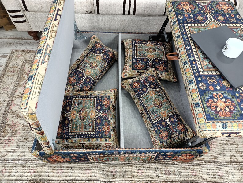 May include: A storage ottoman with a lift-top lid revealing a storage compartment. The ottoman is covered in a colorful patterned fabric with blue, red, and yellow accents. The lid is open and reveals four patterned pillows inside.