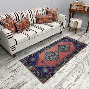May include: A red and blue patterned rug with a white background. The rug is placed in front of a white couch with striped upholstery and pillows. The couch is in a room with whitewashed wood floors.