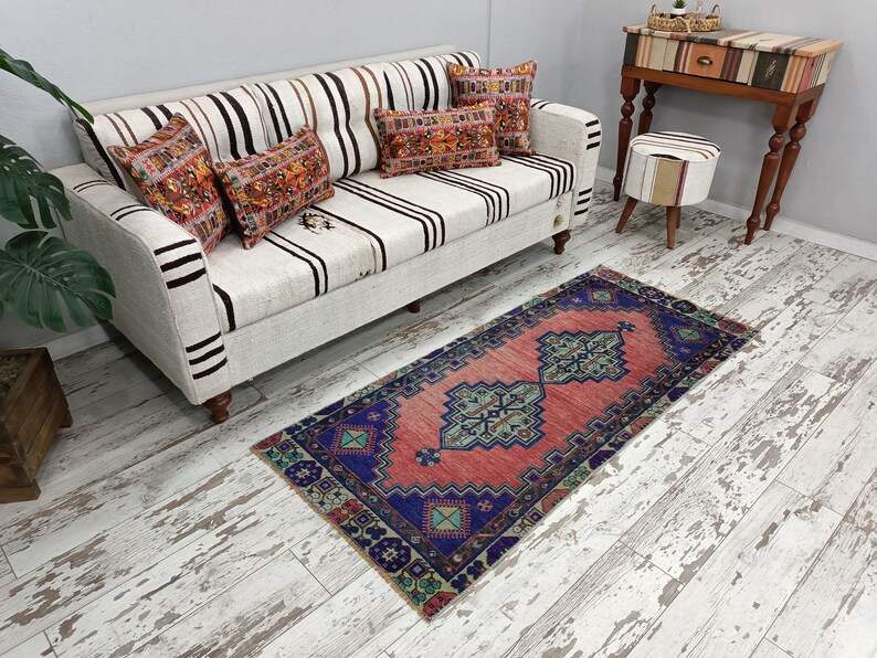 May include: A red and blue patterned rug with a white background. The rug is placed in front of a white couch with striped upholstery and pillows. The couch is in a room with whitewashed wood floors.