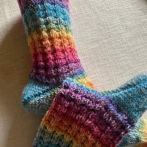 Könnte beinhalten: Ein Paar handgestrickte Socken in einem lebendigen Regenbogen aus Farben. Die Socken haben ein geripptes Bündchen und ein strukturiertes Strickmuster mit Farbtönen von Rot, Orange, Gelb, Grün, Blau und Lila. Die Socken sind aus weichem, möglicherweise Wollgarn.