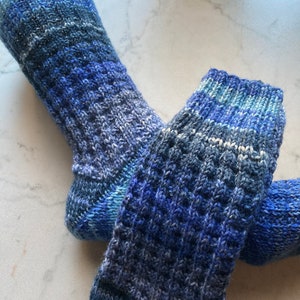 Könnte beinhalten: Ein Paar handgestrickte Socken in Blautönen mit einem strukturierten Muster. Die Socken sind aus weichem Garn gefertigt und wirken warm und gemütlich. Die Farbpalette umfasst verschiedene Blau-, Weiß- und Grautöne.