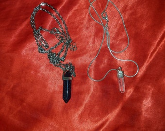 May include: Two necklaces with pendants on a red satin background. One necklace has a silver chain with a dark blue, crystal-like pendant. The other necklace has a silver chain with a clear, crystal-like pendant.