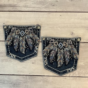 May include: Two dark blue denim pockets with intricate feather embroidery in shades of brown, blue, and silver. The pockets are embellished with small, sparkling rhinestones and feature decorative stitching.