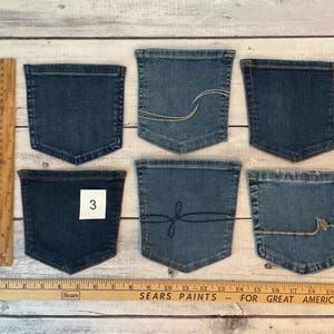 Pode incluir: Seis bolsos de ganga azul dispostos em duas filas de tr&ecirc;s. Os bolsos apresentam v&aacute;rios detalhes de costura, incluindo linhas curvas e um design de la&ccedil;o. Um bolso tem uma etiqueta quadrada branca com o n&uacute;mero 3. Uma r&eacute;gua de madeira mostra medidas em cm.
