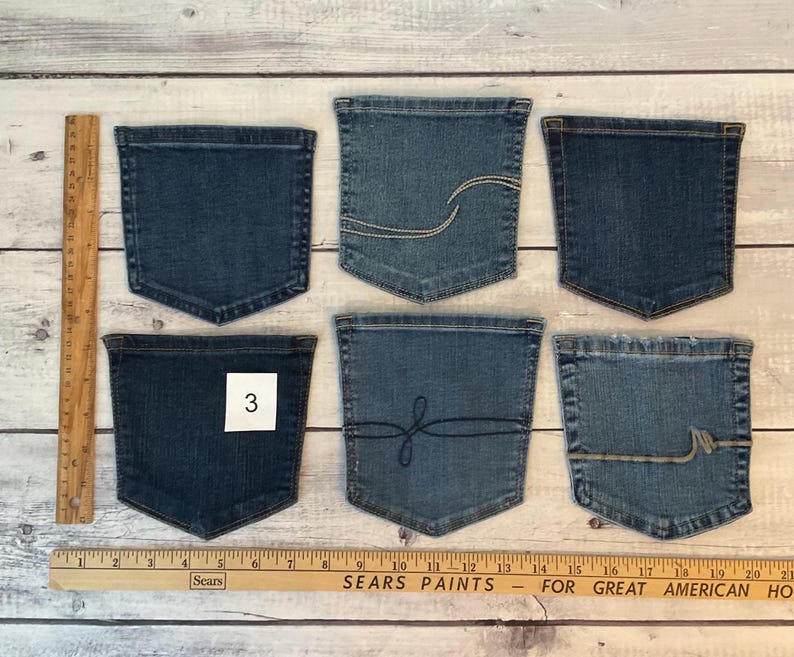 Pode incluir: Seis bolsos de ganga azul dispostos em duas filas de tr&ecirc;s. Os bolsos apresentam v&aacute;rios detalhes de costura, incluindo linhas curvas e um design de la&ccedil;o. Um bolso tem uma etiqueta quadrada branca com o n&uacute;mero 3. Uma r&eacute;gua de madeira mostra medidas em cm.