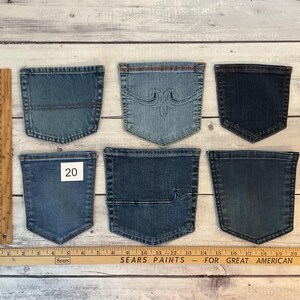 Puede incluir: Seis bolsillos de mezclilla variados en diferentes tonos de azul, dispuestos en dos filas. Un bolsillo presenta costuras decorativas, otro tiene una l&iacute;nea horizontal y uno muestra una etiqueta cuadrada blanca con el n&uacute;mero 20. Se ve una regla de madera.