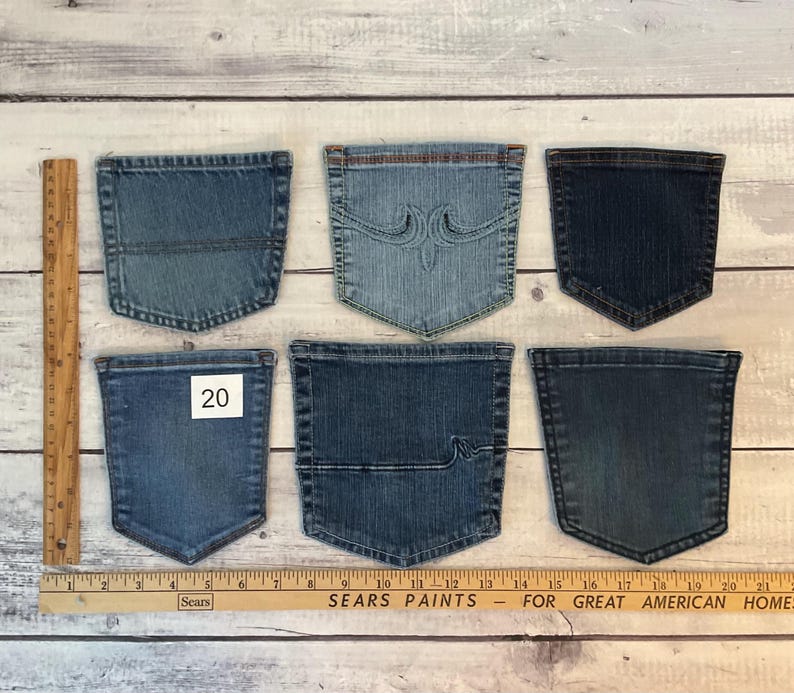 Puede incluir: Seis bolsillos de mezclilla variados en diferentes tonos de azul, dispuestos en dos filas. Un bolsillo presenta costuras decorativas, otro tiene una l&iacute;nea horizontal y uno muestra una etiqueta cuadrada blanca con el n&uacute;mero 20. Se ve una regla de madera.