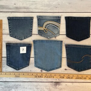 Pode incluir: Seis bolsos de ganga em v&aacute;rios tons de azul, incluindo um com bordado branco e dourado e outro com costura dourada. Uma r&eacute;gua de madeira &eacute; vis&iacute;vel no lado esquerdo e na parte inferior, mostrando medidas at&eacute; 56 cm.