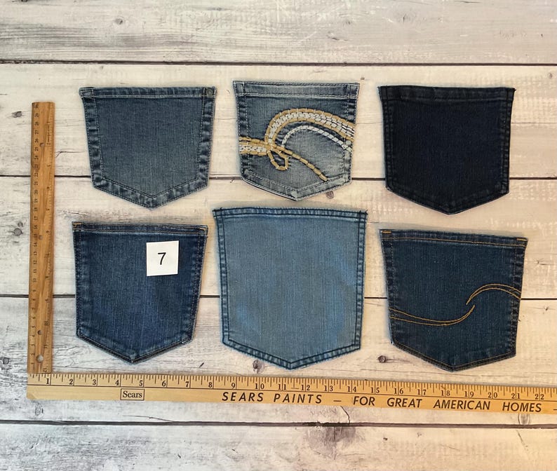Pode incluir: Seis bolsos de ganga em v&aacute;rios tons de azul, incluindo um com bordado branco e dourado e outro com costura dourada. Uma r&eacute;gua de madeira &eacute; vis&iacute;vel no lado esquerdo e na parte inferior, mostrando medidas at&eacute; 56 cm.