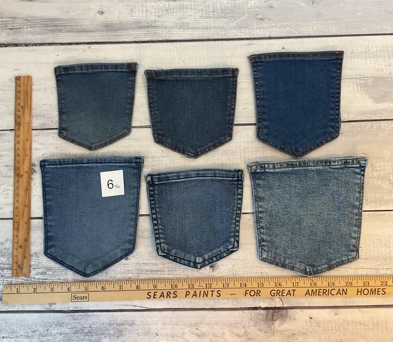Pode incluir: Seis bolsos de ganga azul separados de v&aacute;rios tons e tamanhos dispostos em duas filas. Uma r&eacute;gua de madeira &agrave; esquerda mostra uma altura de cerca de 16,5 cm para o bolso inferior esquerdo, e uma r&eacute;gua horizontal na base mede 56 cm.