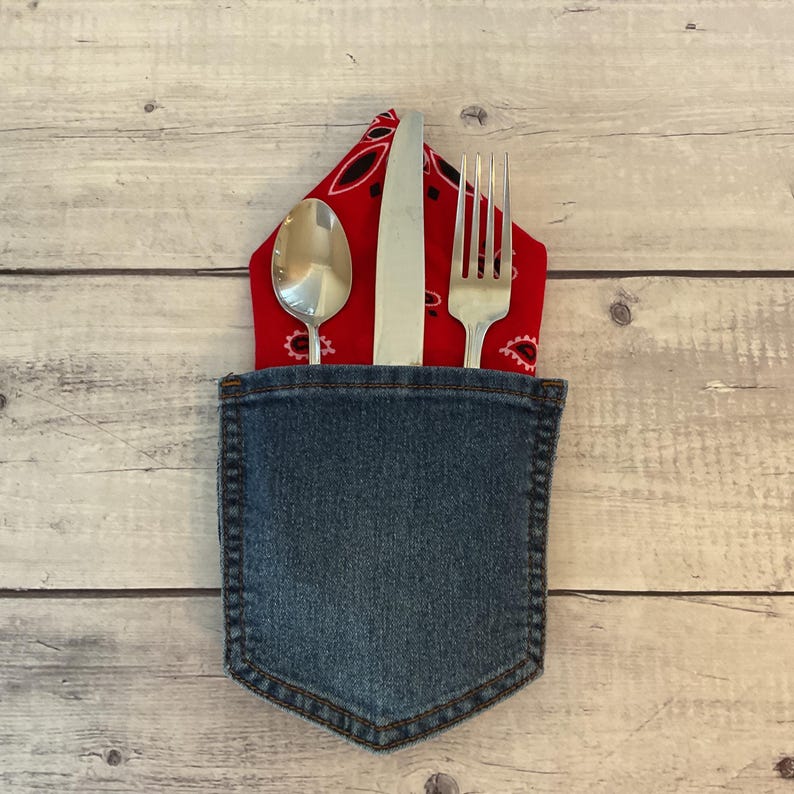 Puede incluir: Un soporte para cubiertos de mezclilla azul que contiene un pa&ntilde;uelo rojo con un patr&oacute;n de cachemira negro. Dentro del bolsillo, una cuchara de metal plateado, un cuchillo de mesa y un tenedor de cuatro dientes est&aacute;n dispuestos verticalmente.