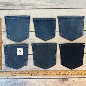 Pode incluir: Seis bolsos de ganga em v&aacute;rios tons de azul dispostos em duas filas, com uma r&eacute;gua de madeira que mostra medidas em cent&iacute;metros no lado esquerdo e na parte inferior. Um bolso apresenta uma etiqueta quadrada branca com o n&uacute;mero 5.