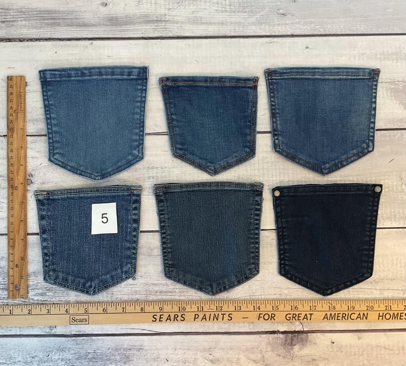 Pode incluir: Seis bolsos de ganga em v&aacute;rios tons de azul dispostos em duas filas, com uma r&eacute;gua de madeira que mostra medidas em cent&iacute;metros no lado esquerdo e na parte inferior. Um bolso apresenta uma etiqueta quadrada branca com o n&uacute;mero 5.