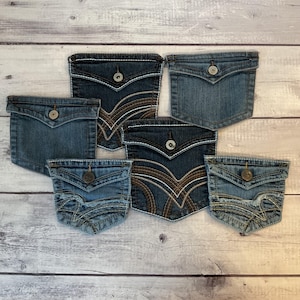 May include: An assortment of denim jean pockets in various shades of blue. Each pocket features decorative stitching in white and brown, with a button closure. The pockets are arranged on a light wood-grain surface.