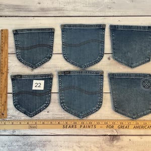Puede incluir: Seis bolsillos de mezclilla azul dispuestos en dos filas, con una regla de madera que muestra una longitud de 53 cm. Los bolsillos tienen detalles de costura, uno tiene una etiqueta Lee, uno un dise&ntilde;o bordado circular y uno una etiqueta con el 22.