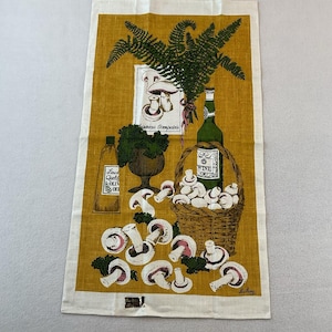 May include: A rectangular tea towel with a mustard yellow background featuring a printed illustration of a wicker basket filled with white mushrooms, a green wine bottle, a bottle of olive oil, a bowl of herbs, and a fern sprig with text.
