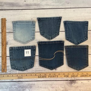 Pode incluir: Seis bolsos de ganga variados em v&aacute;rios tons de azul, variando do claro ao escuro. Um bolso apresenta uma etiqueta quadrada branca com o n&uacute;mero 11 e outro tem costuras decorativas douradas. Uma r&eacute;gua de madeira mostra medidas em cent&iacute;metros.