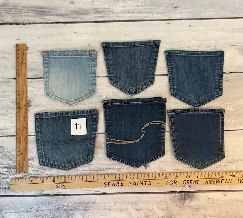 Pode incluir: Seis bolsos de ganga variados em v&aacute;rios tons de azul, variando do claro ao escuro. Um bolso apresenta uma etiqueta quadrada branca com o n&uacute;mero 11 e outro tem costuras decorativas douradas. Uma r&eacute;gua de madeira mostra medidas em cent&iacute;metros.