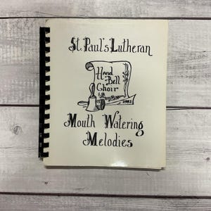 May include: A spiral-bound book with a cream-colored cover. The cover features black calligraphy that reads "St. Paul's Lutheran Hand Bell Choir" and "Mouth Watering Melodies." A bell graphic is also present, with the year 1981.