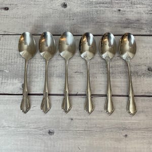 May include: A set of six polished silver-tone metal spoons arranged in a horizontal row. Each spoon features a smooth, oval-shaped bowl and a slender handle that widens toward the base, displaying a subtle decorative embossed floral motif.