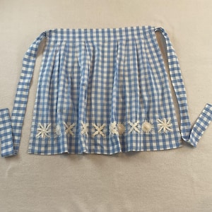 May include: A waist apron featuring a light blue and white gingham check pattern with vertical pleats along the waistband. The bottom hem is decorated with a horizontal row of white embroidered star-shaped motifs and two long fabric ties.