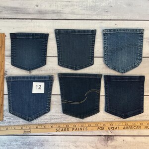 Pode incluir: Seis bolsos de ganga variados em v&aacute;rios tons de azul, dispostos em duas filas. Uma r&eacute;gua de madeira est&aacute; posicionada verticalmente &agrave; esquerda e horizontalmente na parte inferior, mostrando medidas em cent&iacute;metros. Um bolso apresenta uma etiqueta branca com o n&uacute;mero 12.