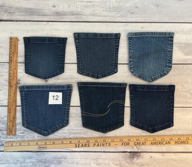 Pode incluir: Seis bolsos de ganga variados em v&aacute;rios tons de azul, dispostos em duas filas. Uma r&eacute;gua de madeira est&aacute; posicionada verticalmente &agrave; esquerda e horizontalmente na parte inferior, mostrando medidas em cent&iacute;metros. Um bolso apresenta uma etiqueta branca com o n&uacute;mero 12.
