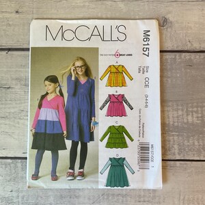 May include: A McCall's M6157 sewing pattern envelope for children's dresses in sizes 3-4-5-6. The front displays illustrations of four dress variations with long sleeves and wrap-style bodices, alongside photos of two children wearing the finished garments.