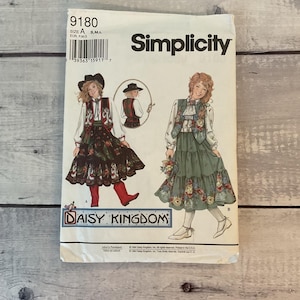 May include: A Simplicity 9180 Daisy Kingdom sewing pattern envelope featuring illustrations of two outfits. Outfit A includes a dark skirt with floral patterns, a vest, and a white blouse. Outfit B shows a green tiered skirt with floral trim and a vest.
