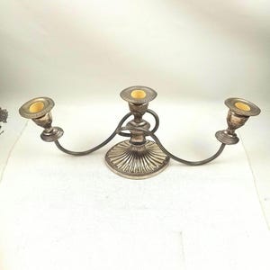 May include: A silver-toned metal candelabra featuring three candle holders with cream-coloured inserts. The central base is circular with a fluted design, and two curved arms extend outward to support the side candle holders, all with decorative rims.
