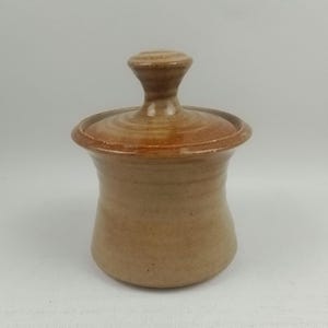 Peut inclure: Un petit pot en céramique fait à la main avec un couvercle. Le pot est de couleur brun clair avec un glaçage brun légèrement plus foncé. Le couvercle a un bouton sur le dessus.