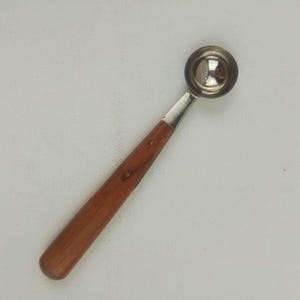 May include: A stainless steel melon baller with a wooden handle. The scoop is a shiny silver color, and the handle is a warm brown with a smooth finish. The tool is designed for scooping melon or other soft fruits.