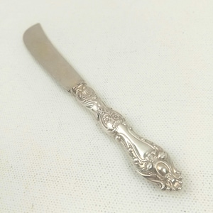 May include: A silver butter knife with a curved blade and an ornate, floral-patterned handle. The knife is made of a shiny, reflective metal and is designed for spreading butter or other soft spreads. The handle features detailed scrollwork.