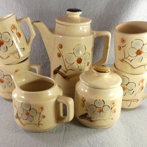 May include: A vintage ceramic tea set with a floral design. The set includes a teapot, creamer, sugar bowl, and four mugs. The pieces are beige with brown and blue floral accents.