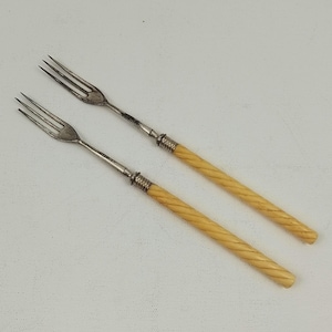 May include: Two antique forks with silver tines and cream-coloured, twisted handles. The forks have a classic design, with a decorative silver band where the handle meets the tines. The handles have a spiral pattern.