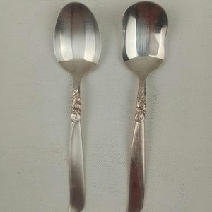 May include: Two silver spoons with decorative floral patterns on the handles. One spoon has a rounded bowl, and the other has a squared-off bowl. The spoons are set against a light background.