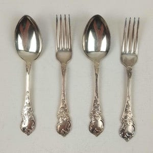 May include: Four pieces of silver cutlery: two spoons and two forks. The spoons have oval bowls, and all pieces feature ornate, floral-style detailing on the handles. The silverware is arranged on a white surface.