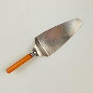 May include: A vintage cake server. It has a stainless steel blade and an orange-brown handle. The handle is cylindrical and appears to be made of a smooth material. The server is designed for cutting and serving cakes, a classic kitchen utensil.