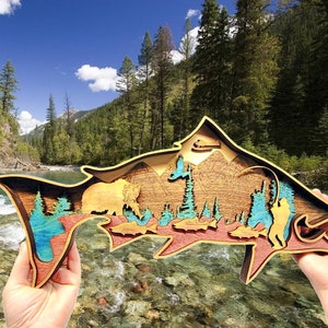 May include: A wooden wall art sculpture of a salmon with a mountain scene inside. The salmon is painted in shades of blue, green, and brown. The mountain scene is painted in shades of brown and green. The sculpture is made of wood and is approximately 12 inches long.