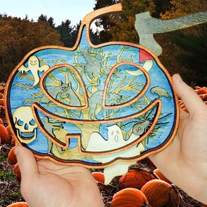 May include: A wooden pumpkin-shaped Halloween decoration with a blue and green painted design. The decoration features a skull, ghosts, bats, and a tree with branches. The decoration is being held in front of a field of pumpkins.