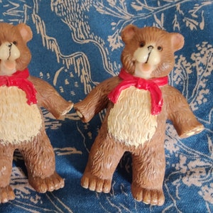 May include: Two brown teddy bear ornaments with cream-coloured bellies and red scarves. The bears are holding hands and have smiling faces. The background is a blue patterned fabric.
