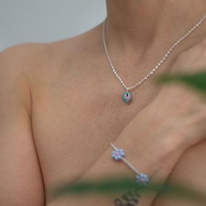 Könnte beinhalten: Eine silberne Kettenkette mit einem blauen und rosafarbenen Augenanhänger. Die Kette wird von einer Person mit einem Blumenarmband am Handgelenk getragen.
