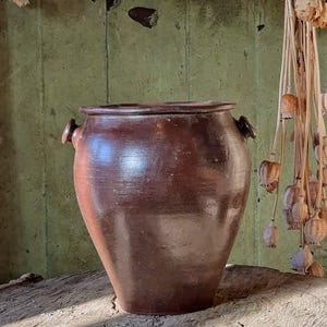 May include: A brown ceramic pot with two small handles, set on a rustic wooden table. The pot has a rounded body and a slightly flared rim. Dried flowers hang in the background, adding a vintage aesthetic.